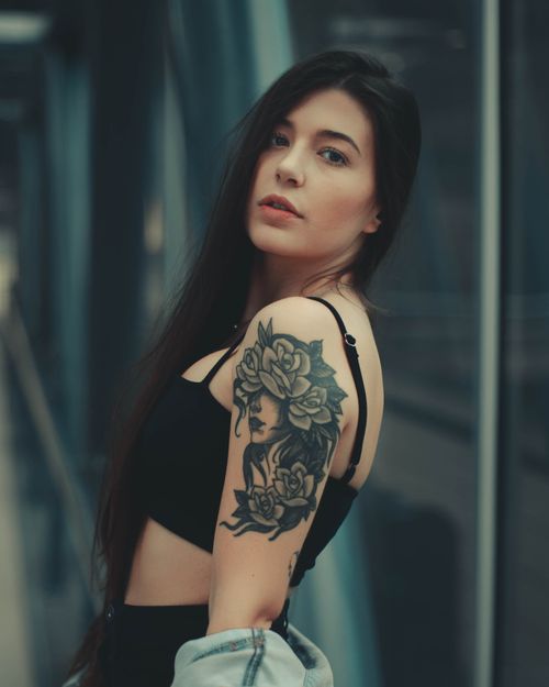 Free A woman with an artistic arm tattoo poses confidently, looking at the camera indoors. Stock Photo