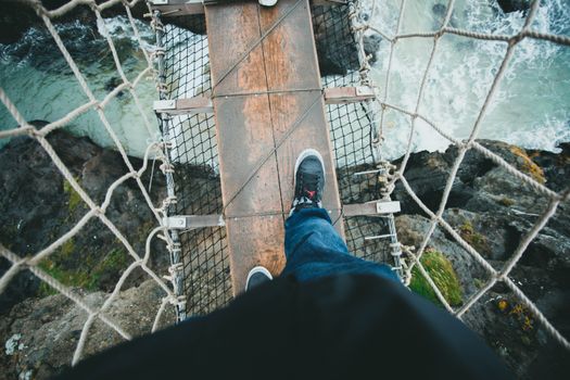 POV shot of a person walking across a wooden rope bridge over a flowing river.