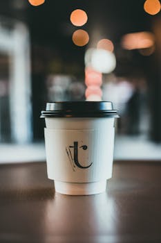 A close-up of a coffee cup with a branded sleeve on a table, set against a blurred background with bokeh lighting.
