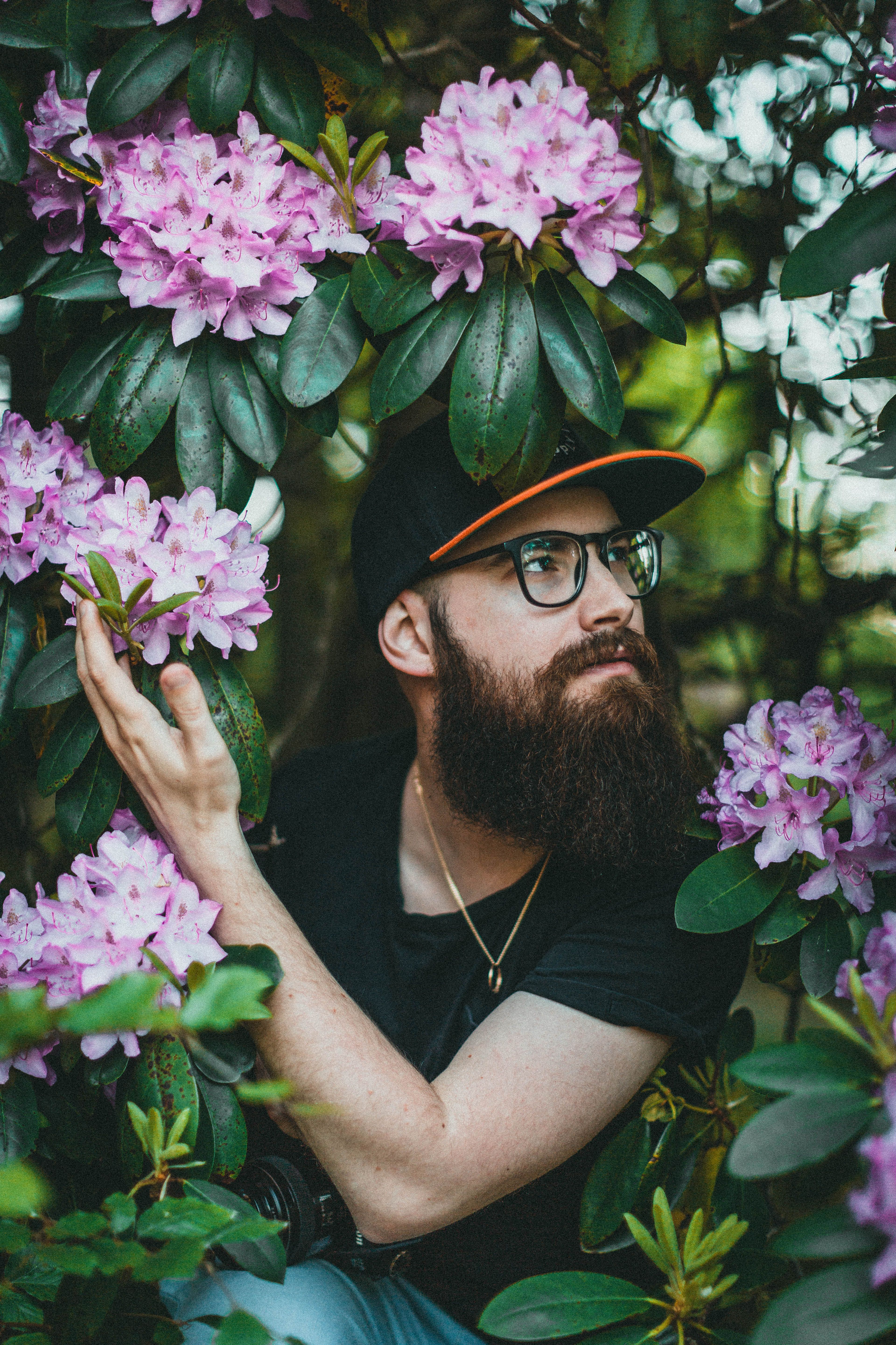 Bearded man wearing glasses and cap amidst pink rhododendrons in garden.