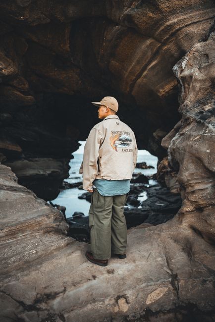 Man wearing a fishing-themed jacket explores a rocky coastal cave, capturing adventure and style.
