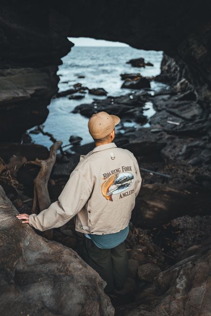 Back view of a man in a stylish jacket standing at a cave's entrance overlooking the sea.