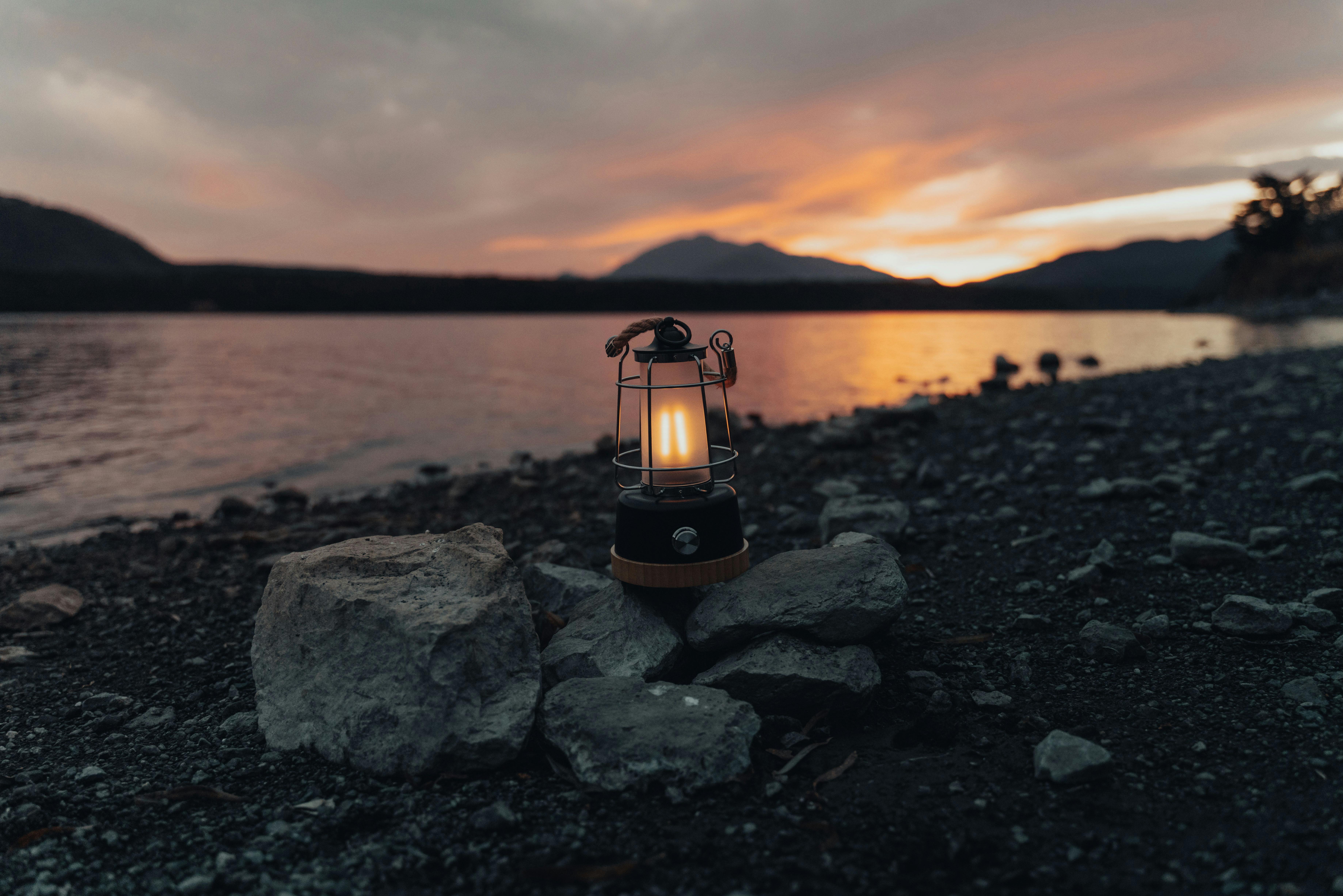 A rustic lantern illuminates a tranquil rocky shore by a lake at sunset in Japan.