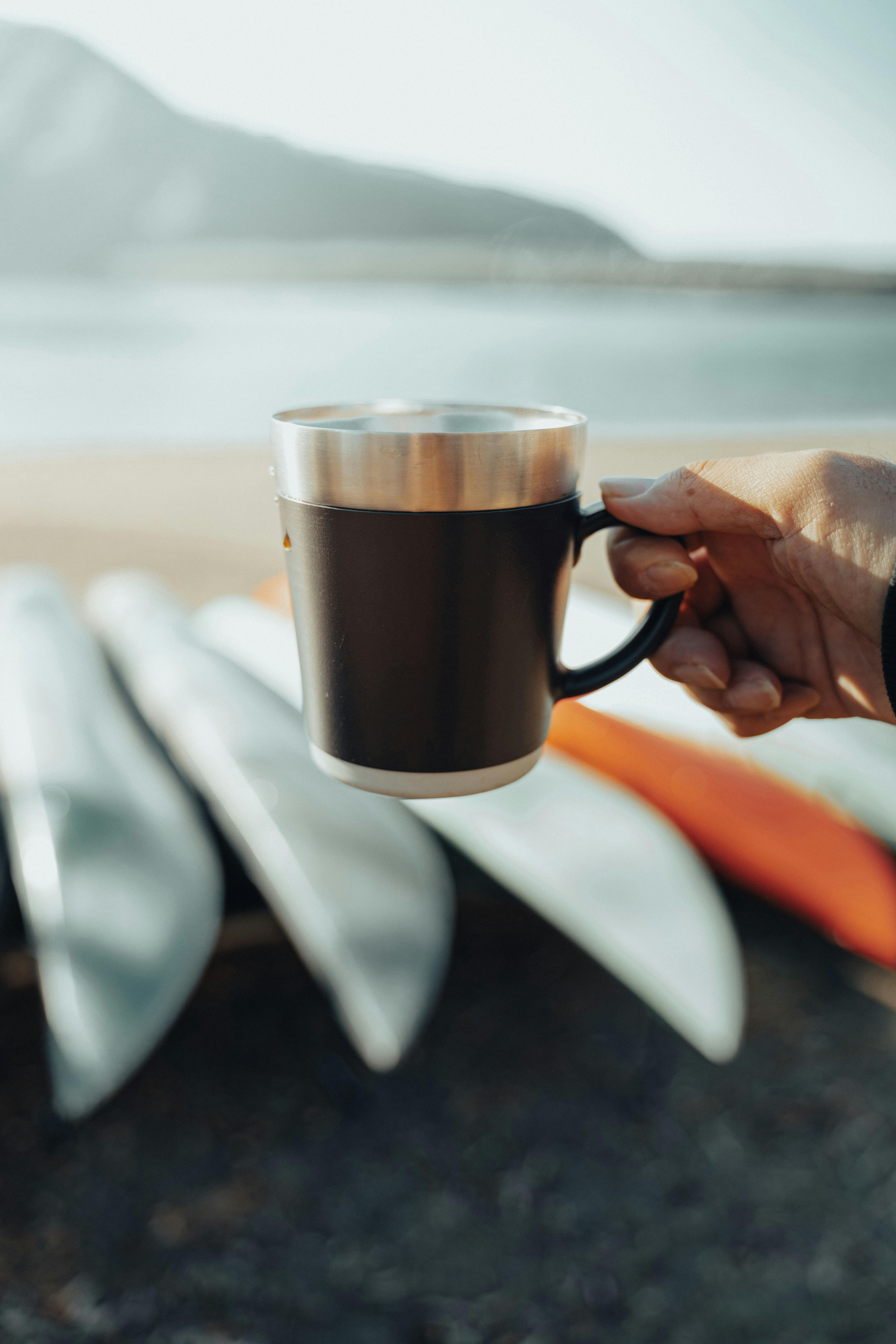 Hand holding a cup by a lake with kayaks in the background, creating a serene outdoor vibe.