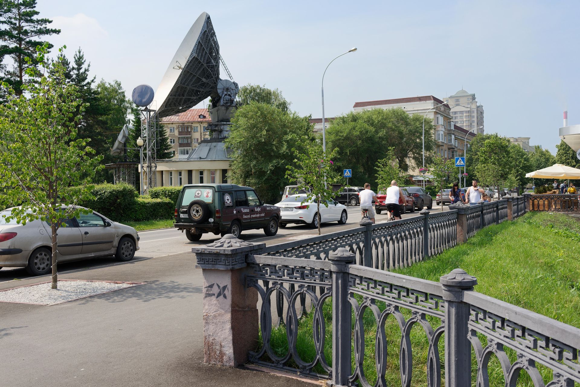 A busy urban street scene featuring a large satellite dish, cars, and pedestrians.