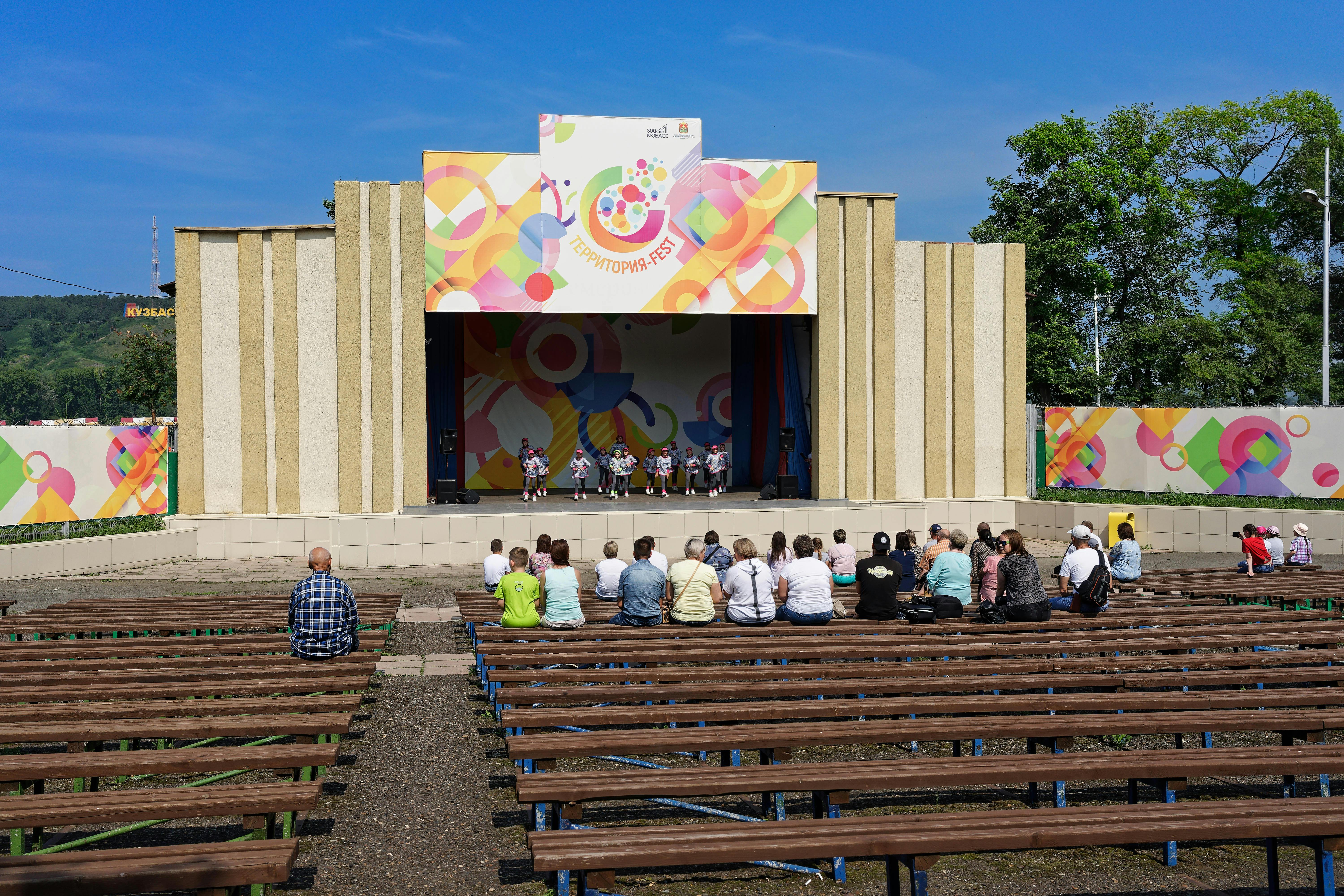 Free Colorful festival stage with audience seated on wooden benches enjoying a daytime performance. Stock Photo
