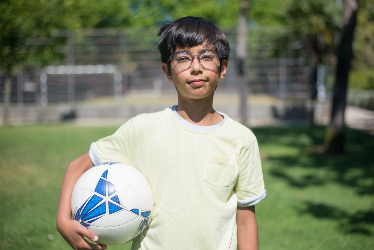 young boy with glasses holding a soccer ball in a sunny park in portug