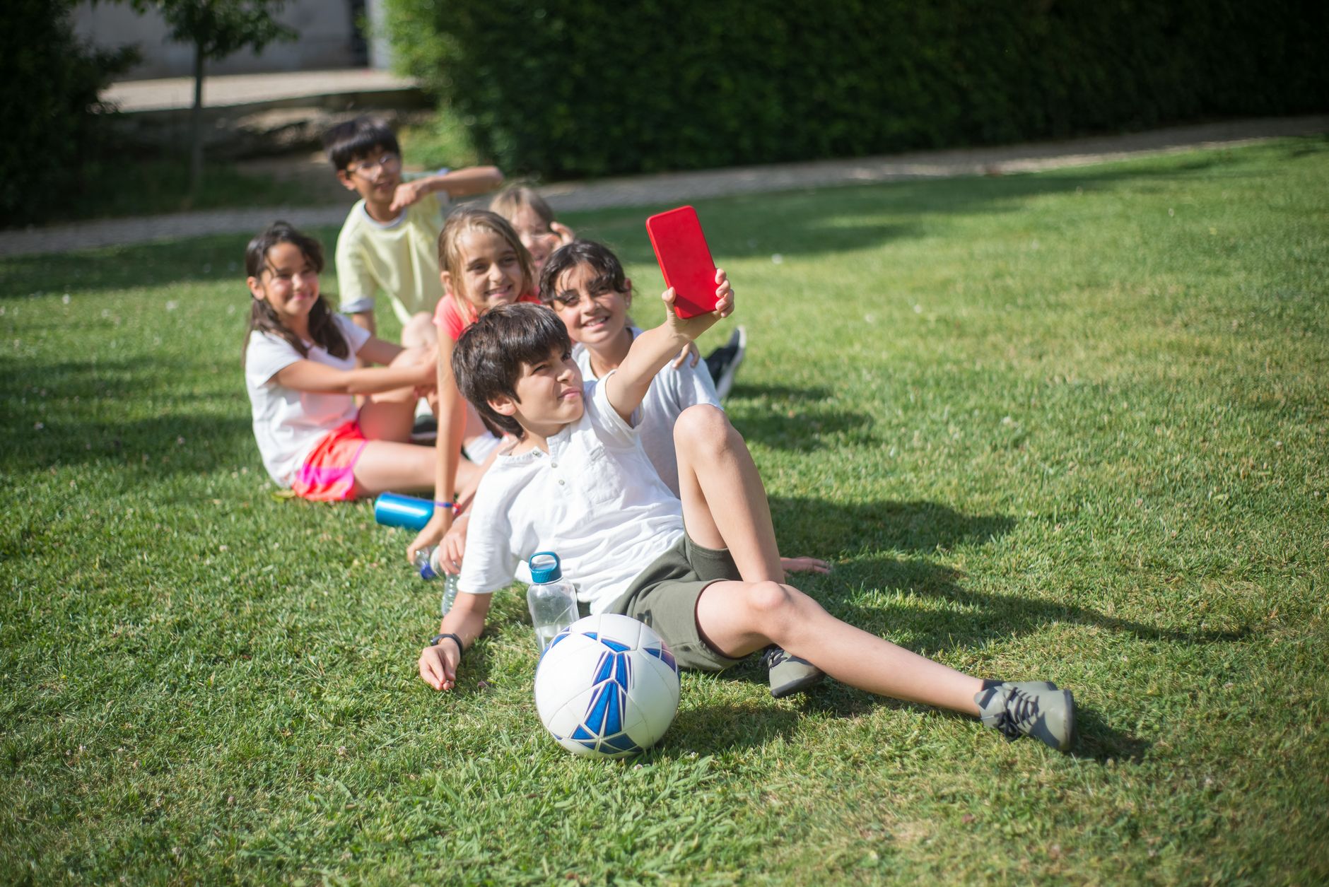 A joyful group of kids sitting on a soccer field, capturing a selfie moment outdoors in daylight.