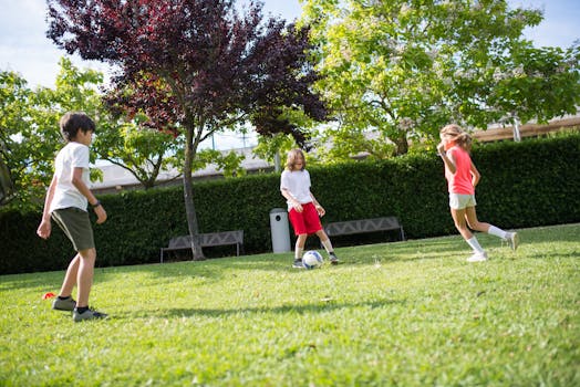 Kids playing football on a grassy field surrounded by green trees, enjoying a sunny day.