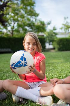 A young girl smiling while holding a soccer ball in a sunny park, sitting on the grass.