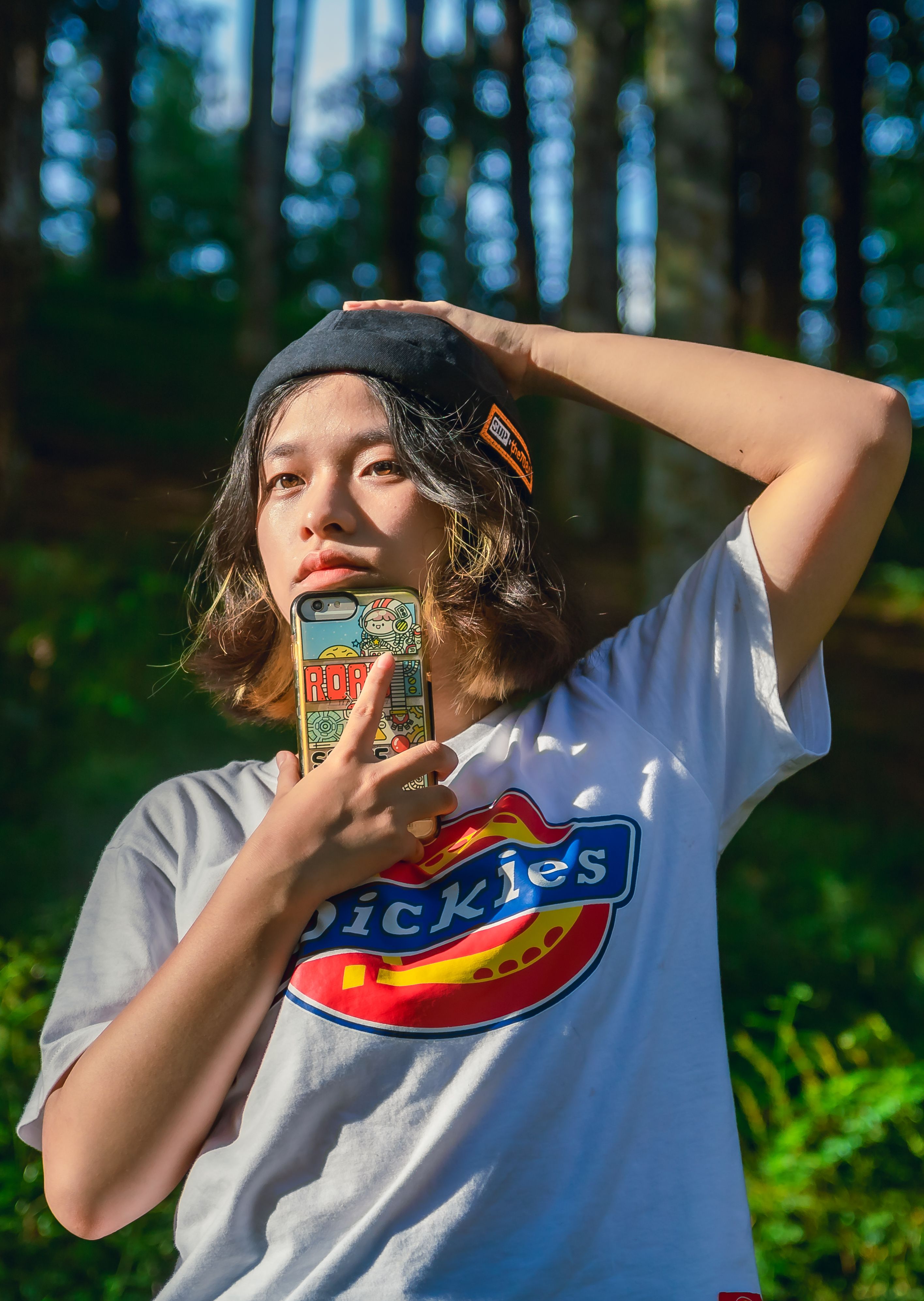 Vibrant portrait of a young woman in a forest holding a smartphone; sunny day, casual attire.