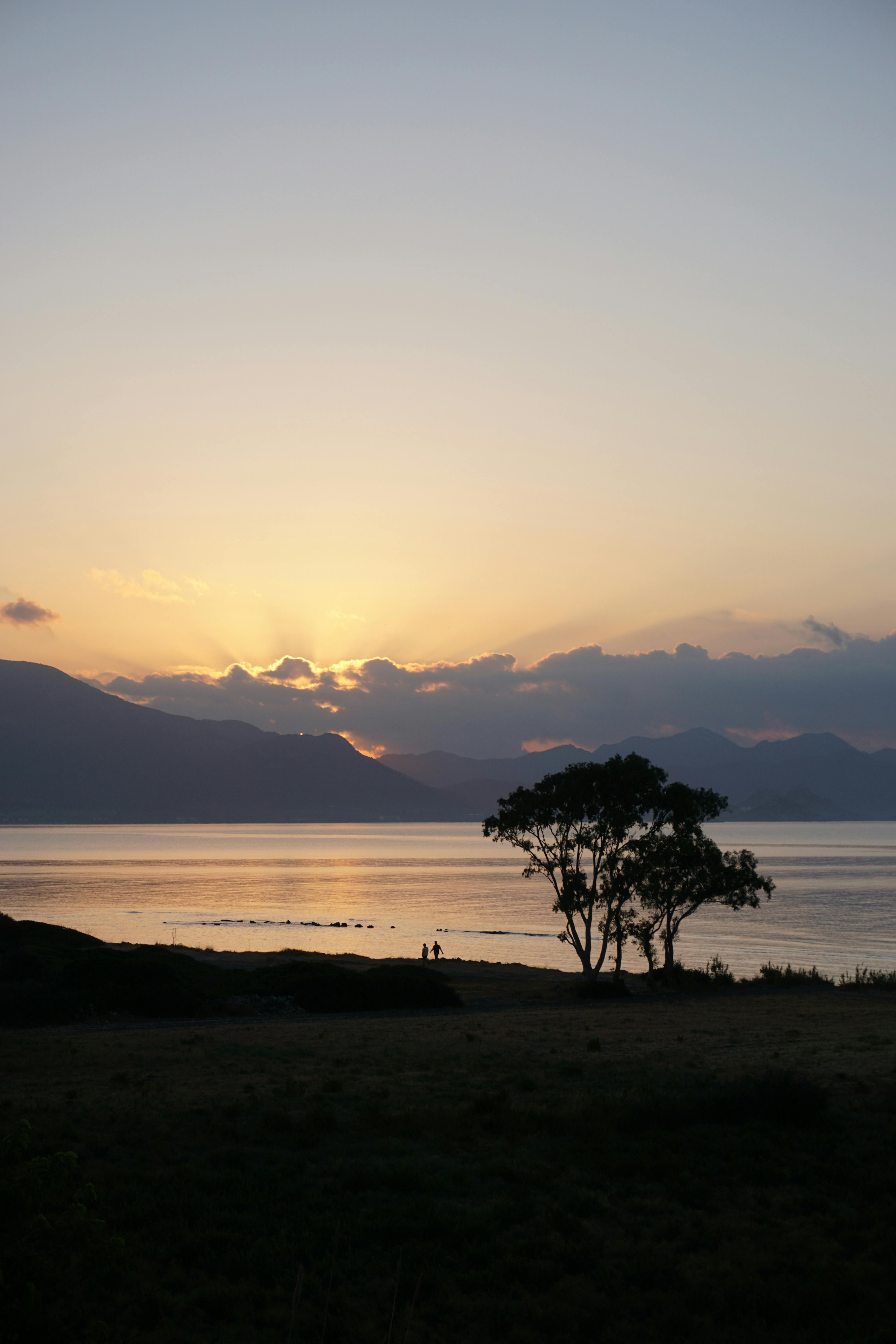 A tranquil sunset scene with silhouetted trees by a mountain-framed lake.