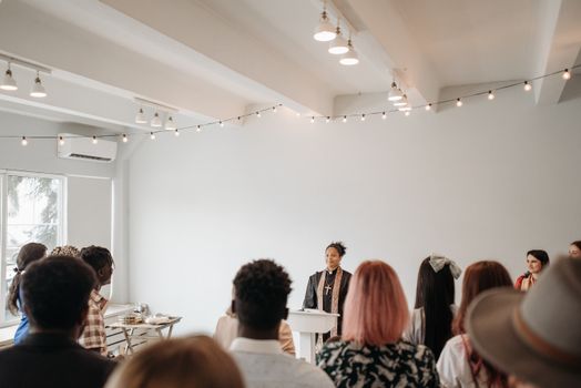 A diverse group of people listens to a pastor in a well-lit church with string lights.