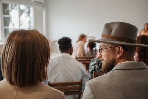Free A man wearing a hat and glasses sits among attendees at an indoor seminar. Stock Photo