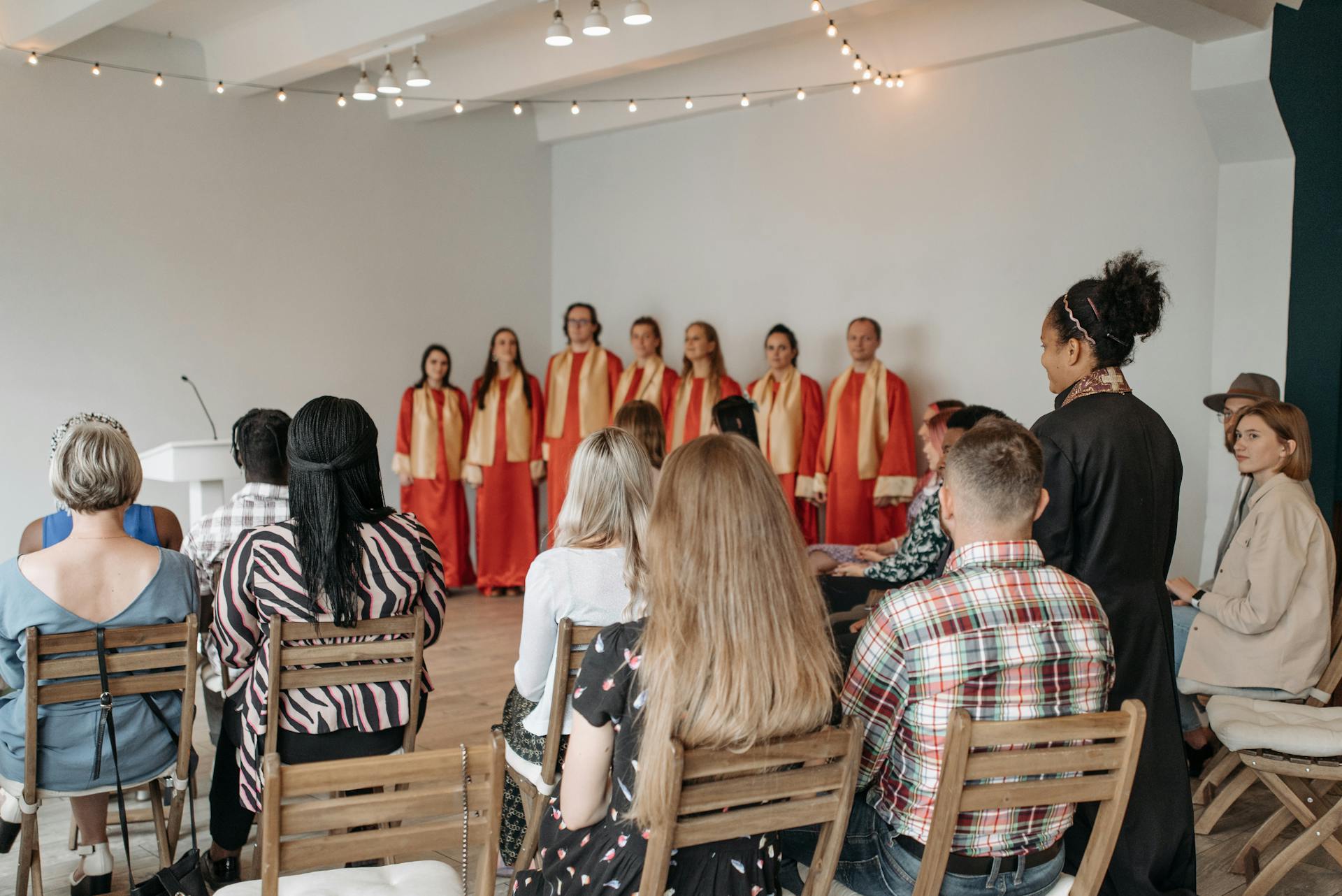 A choir in red robes performing indoors with a seated audience in a community event setting.