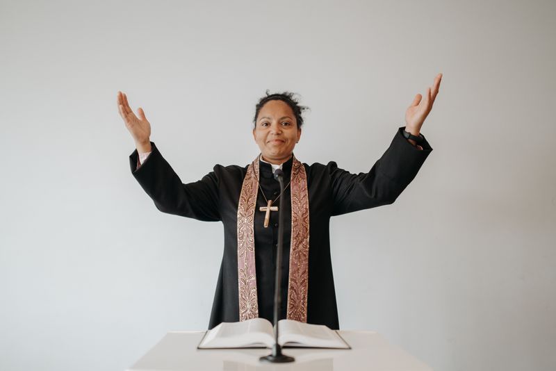 A Black woman pastor in a black clerical robe with an embroidered stole and gold cross stands at a wooden lectern holding an open Bible, hands raised in mid-sermon against a clean white background.