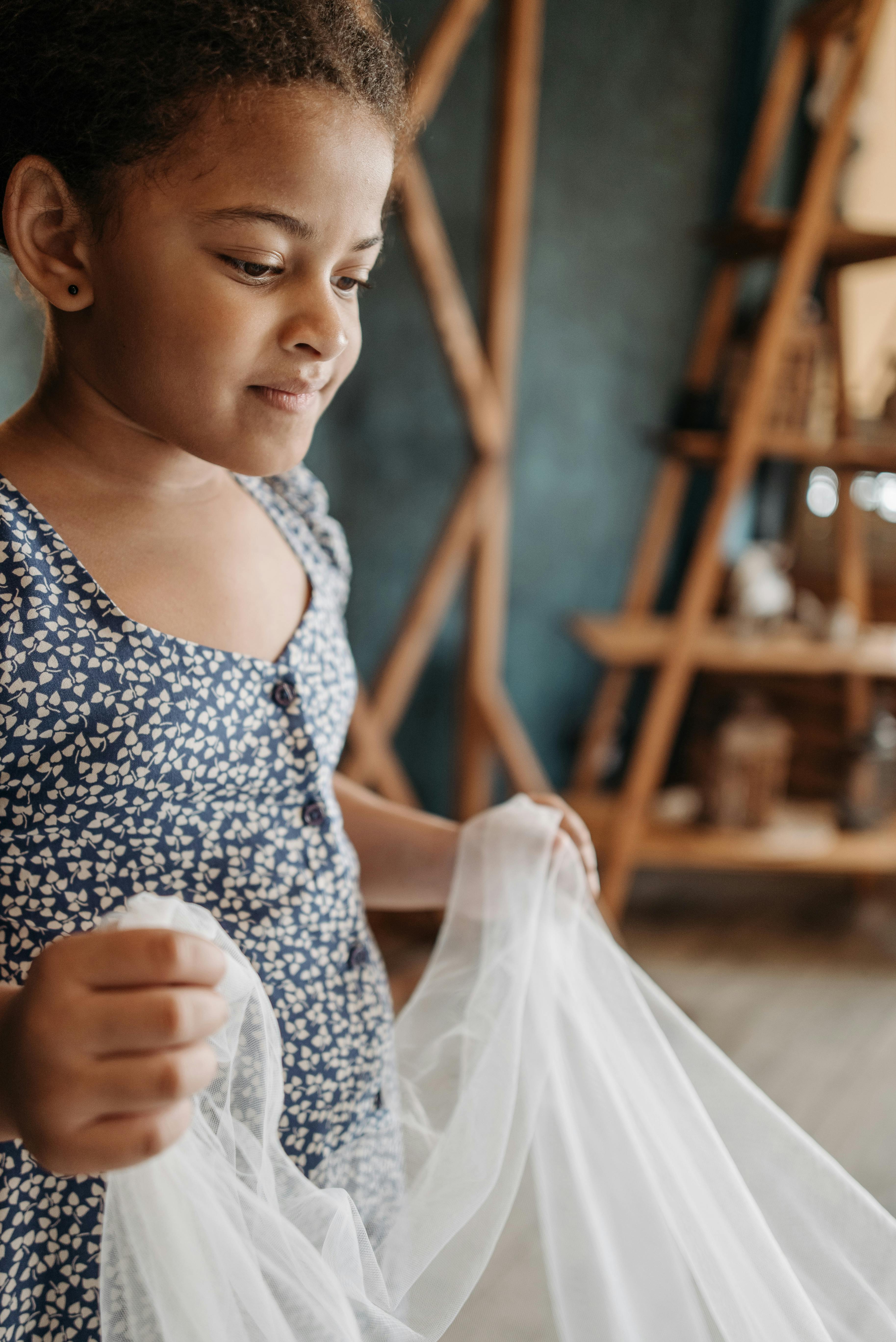 A young girl in a floral dress holds white fabric in a cozy indoor setting.