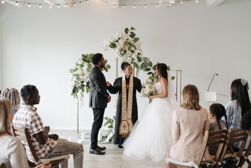 Free An indoor wedding ceremony featuring a bride, groom, and female officiant with diverse guests. Stock Photo