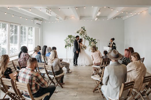 A small gathering witnesses a beautiful wedding ceremony indoors with wooden chairs and floral decor.