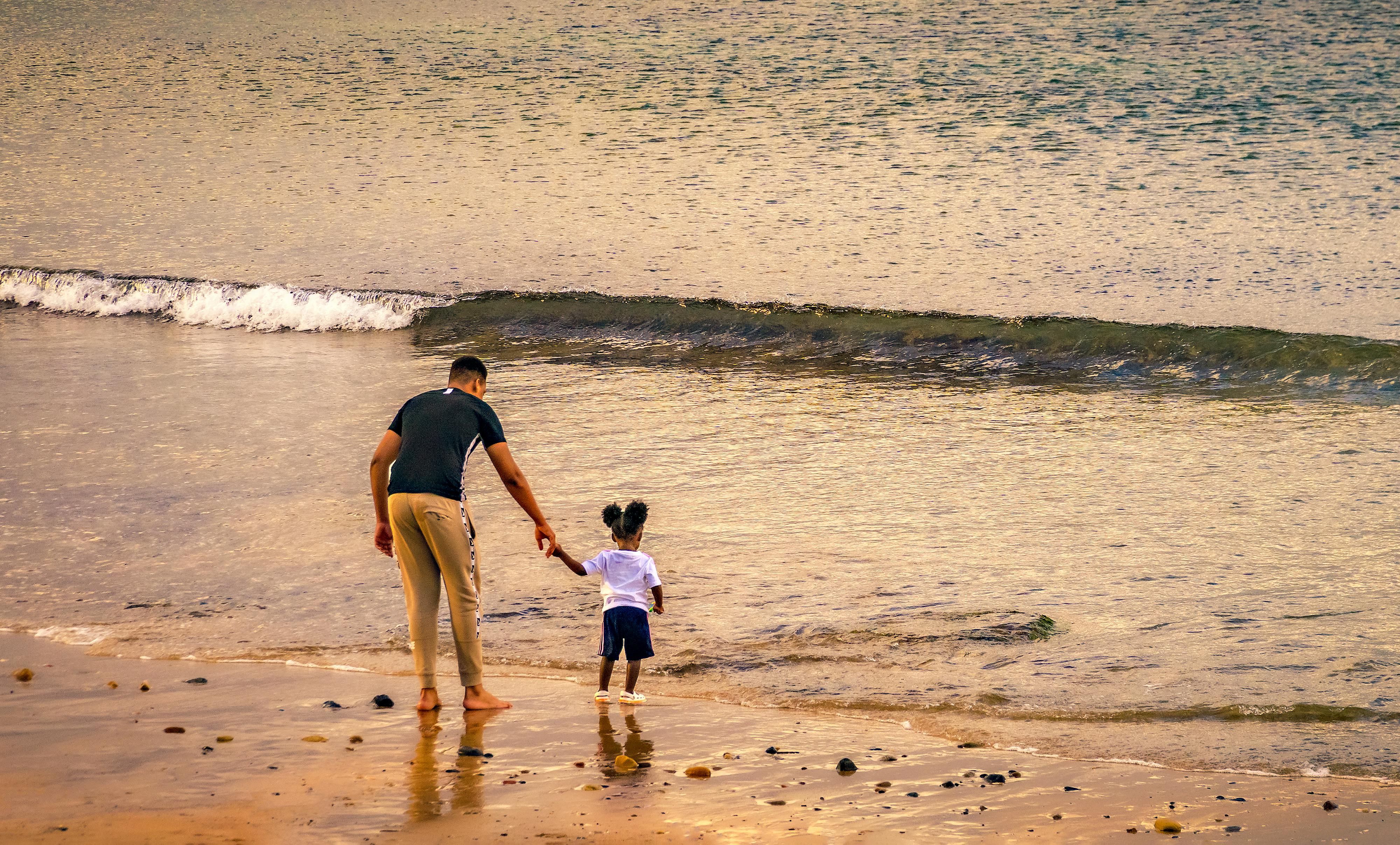 A father and child holding hands while walking along a sandy beach near ocean waves