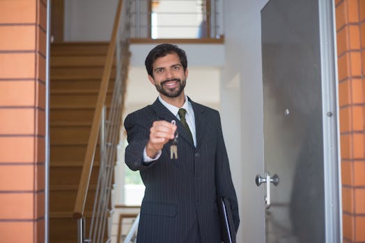 Smiling realtor in a black suit handing keys through an open door inside a modern house.
