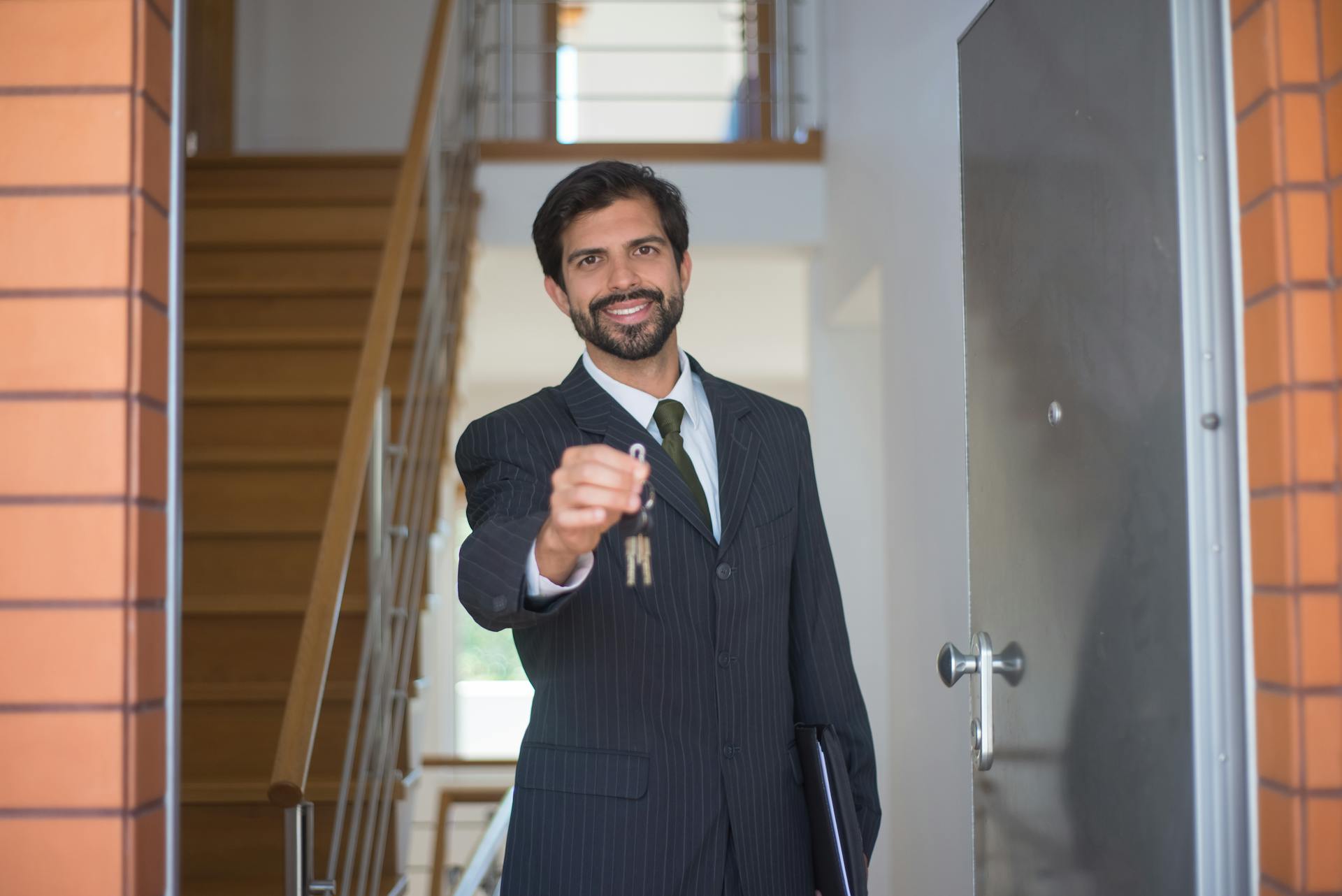 Smiling realtor in a black suit handing keys through an open door inside a modern house.