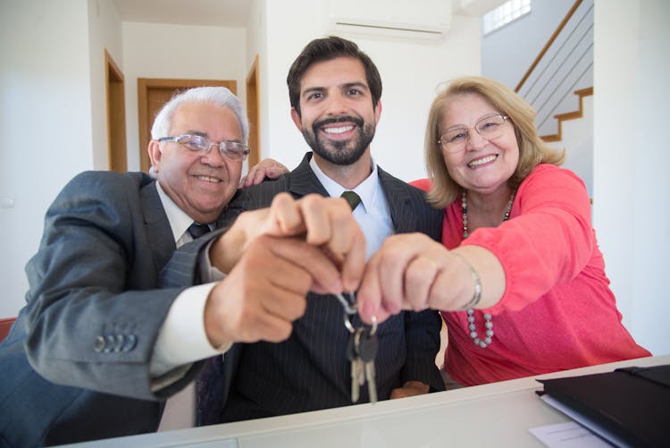 Elderly Couple Holding Keys Of A House Besi8de A Man In Black Suit Jacket