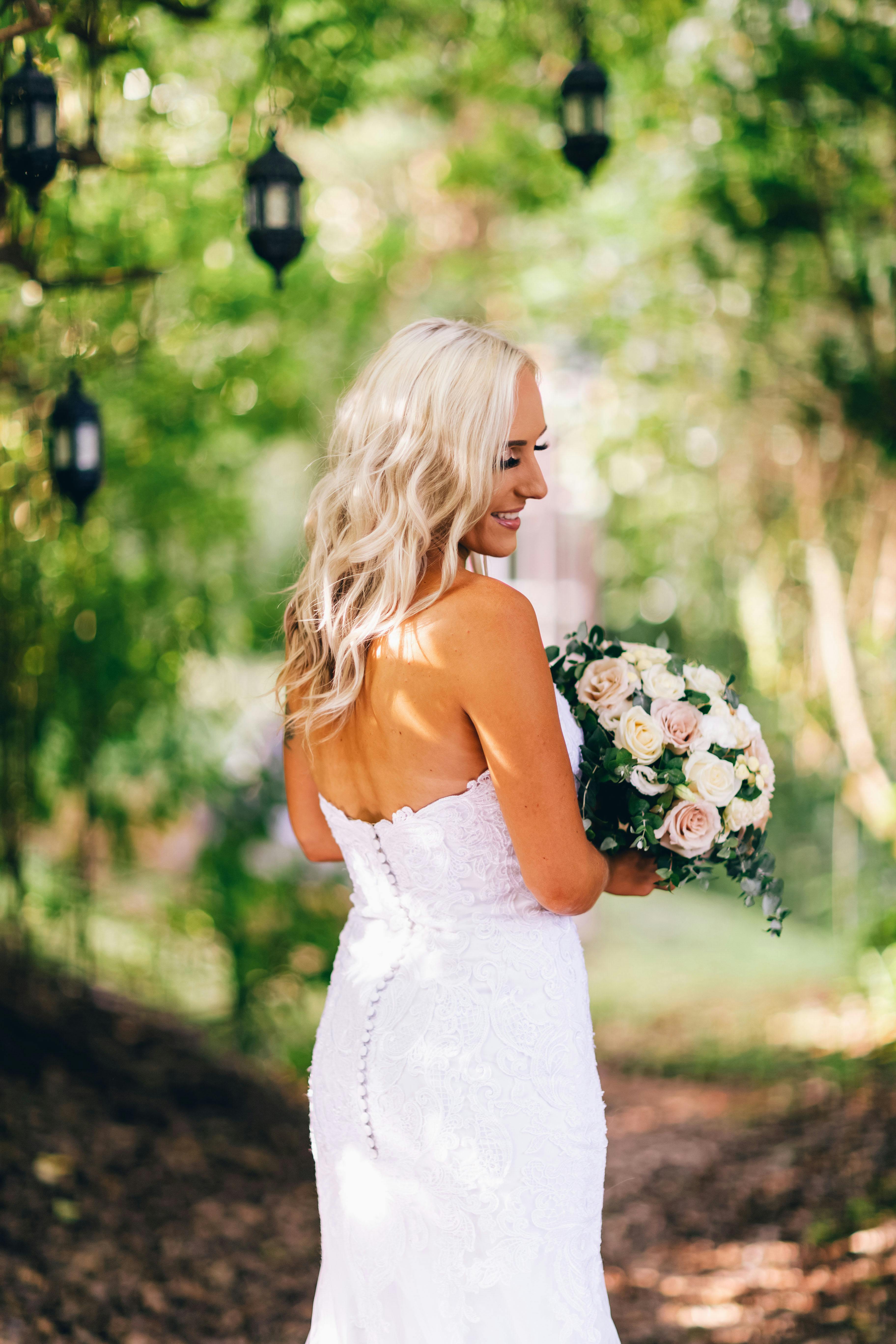 Blonde bride holding bouquet in sunny outdoor setting with lush greenery.
