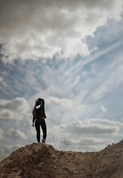 Free Woman in Black Long Sleeve Shirt Standing on Brown Rock Under White Clouds and Blue Sky Stock Photo