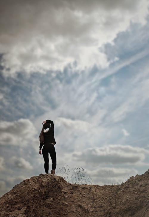 Woman in Black Long Sleeve Shirt Standing on Brown Rock Under White Clouds and Blue Sky