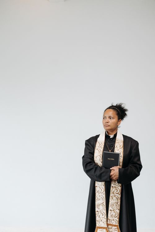 Free A female pastor standing indoors holding a Bible, wearing a cassock and stole. Stock Photo