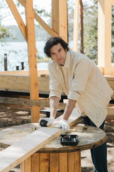 A male carpenter measures wood outdoors at a construction site by the lake.