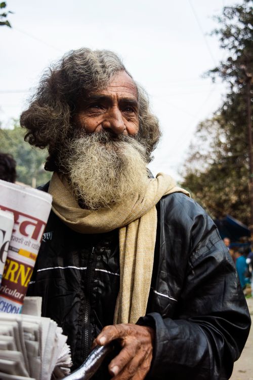Free Elderly man with a large beard holding newspapers outdoors, wearing a scarf and black leather jacket. Stock Photo