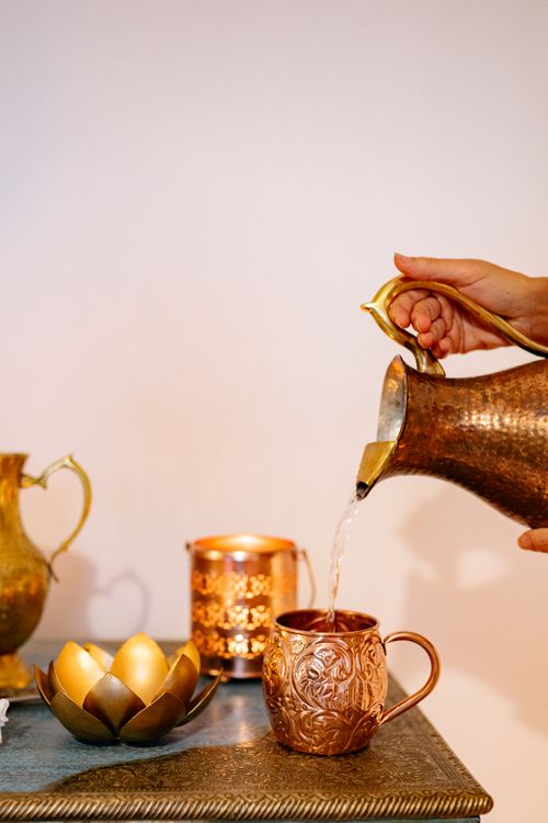 Free A graceful bronze pitcher is seen pouring water into a decorative mug on a table, showcasing elegant design. Stock Photo