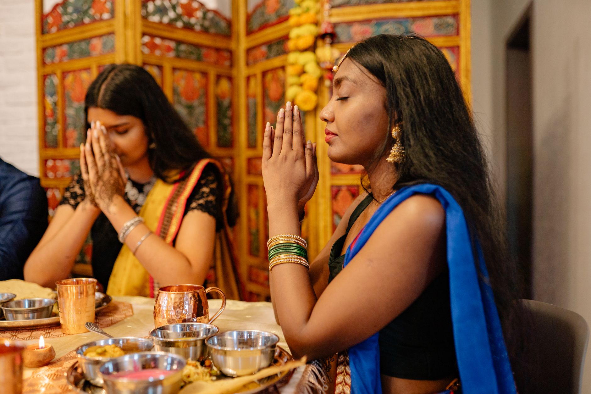 Two Indian women praying during Diwali festival, adorned in traditional attire with a festive setting.