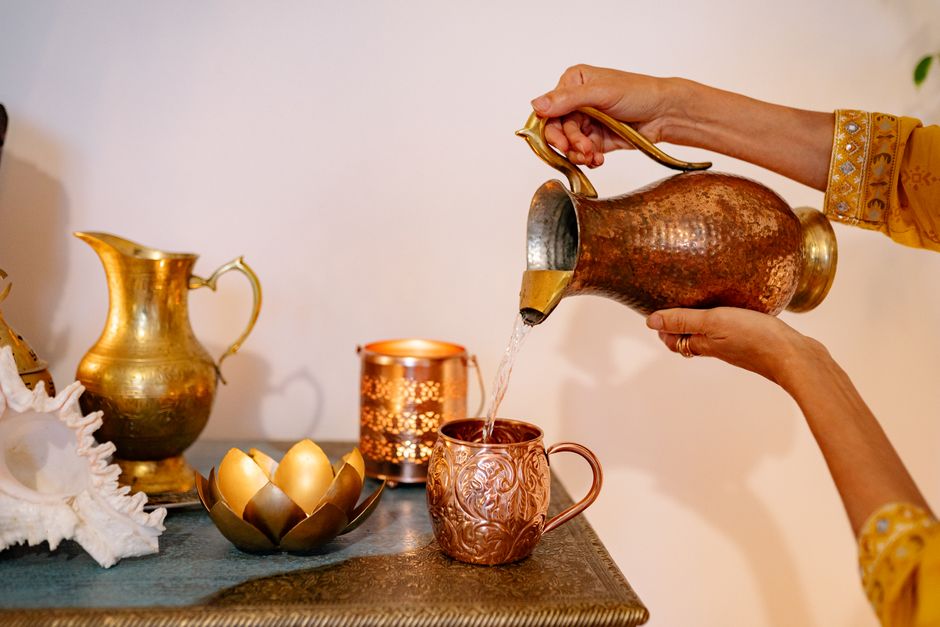 A person pours water from a decorative pitcher into a copper mug surrounded by festive decor.