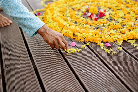A person's hand arranges colorful marigold petals into a flower design on a wooden floor, celebrating Diwali.