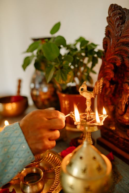 Free A close-up of a hand lighting a brass oil lamp during Diwali with a Ganesha statue and potted plants. Stock Photo
