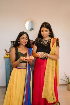 Two South Asian women in traditional attire celebrating Diwali with lighted diyas indoors.