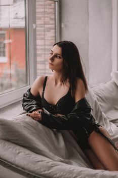 Woman in lingerie with a leather jacket posing thoughtfully by a window indoors.