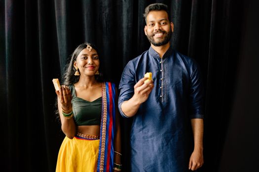A cheerful couple dressed in traditional Indian attire celebrating Diwali with sweets indoors.