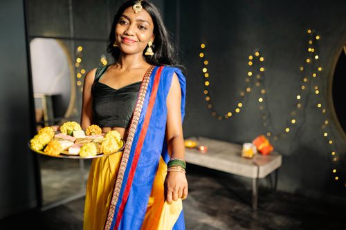 Free Smiling Indian woman in traditional attire holding a festive food platter indoors with decorative lights. Stock Photo