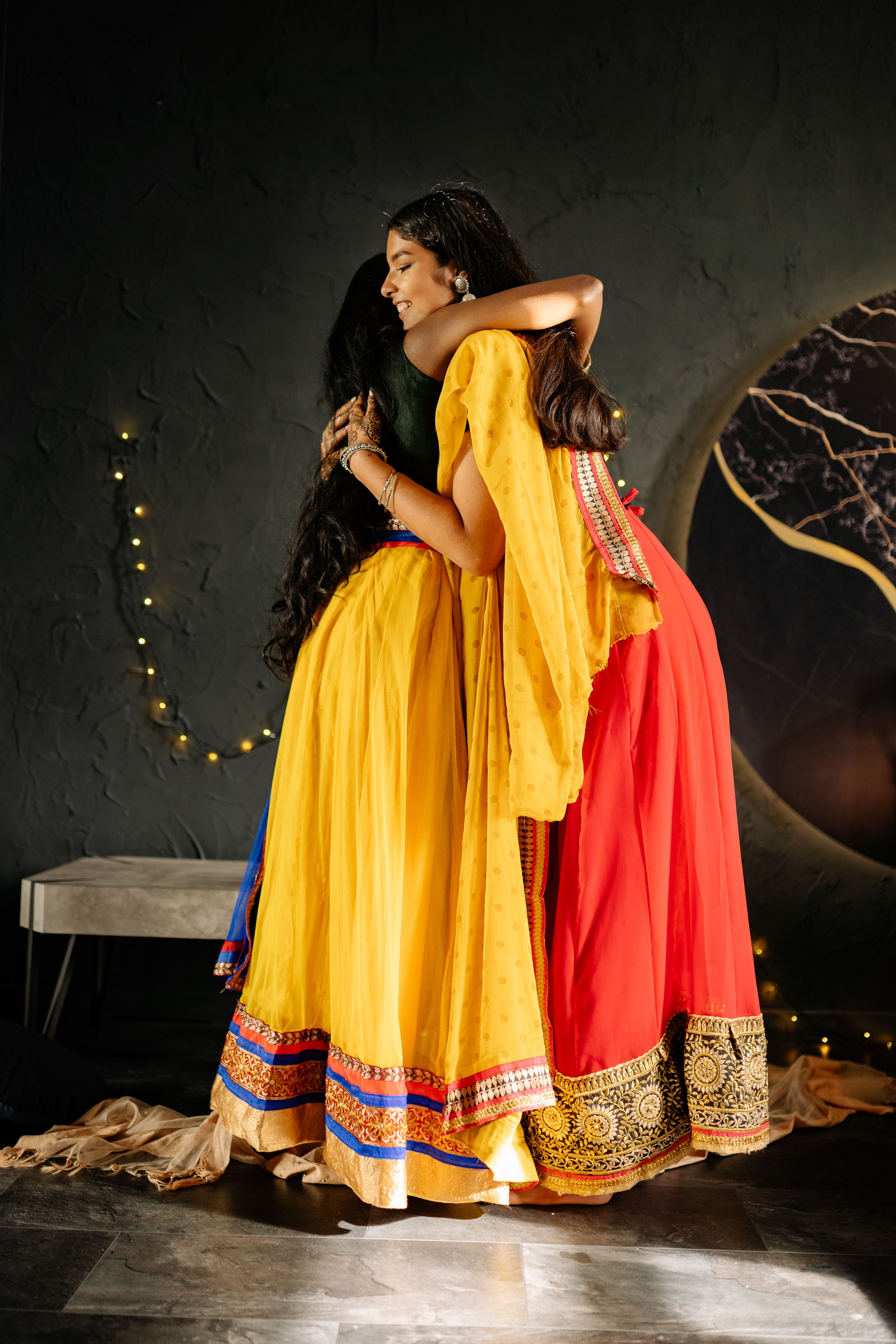 Two women warmly embrace in vibrant traditional dresses, celebrating indoors. Perfect for festive cultural themes.