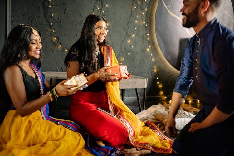 Women Wearing Sari Holding Presents While Sitting In Front Of A Man Wearing Sherwani