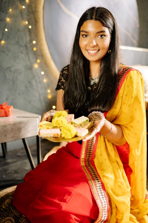 Free Smiling South Asian woman in sari holding a plate of Diwali sweets indoors with festive lights. Stock Photo
