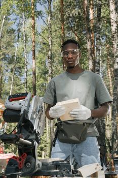 African American carpenter holding wood plank with circular saw in a forest setting.