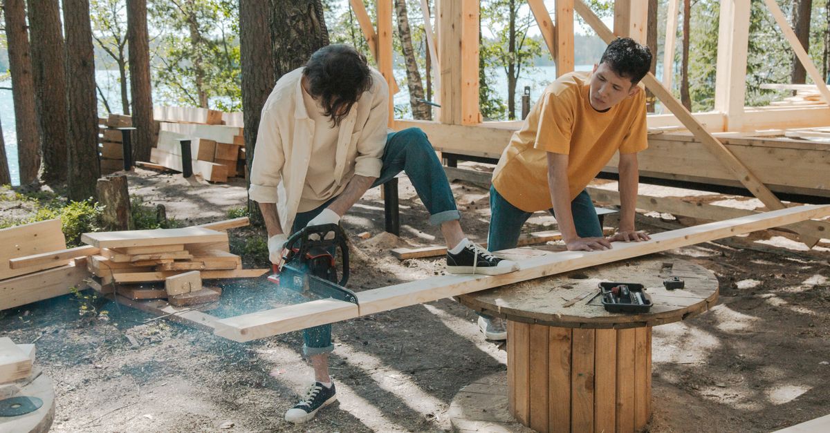 Two adult men working on wood construction project outdoors, using a chainsaw.