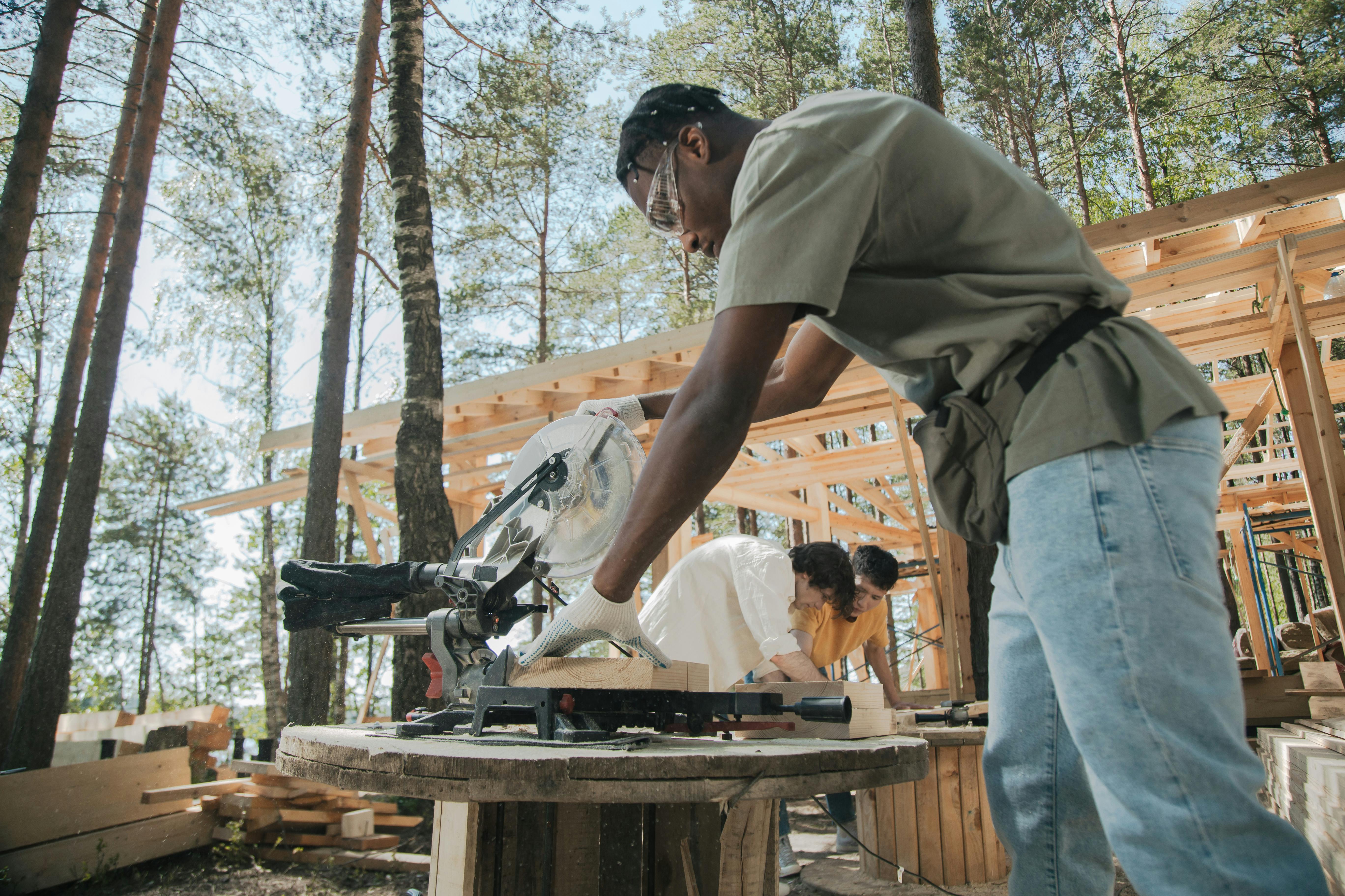 Workers on a construction site cutting wood planks with a circular saw outdoors.