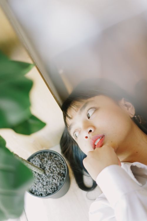 Free A young woman lies thoughtfully beside an indoor plant, evoking a serene mood. Stock Photo
