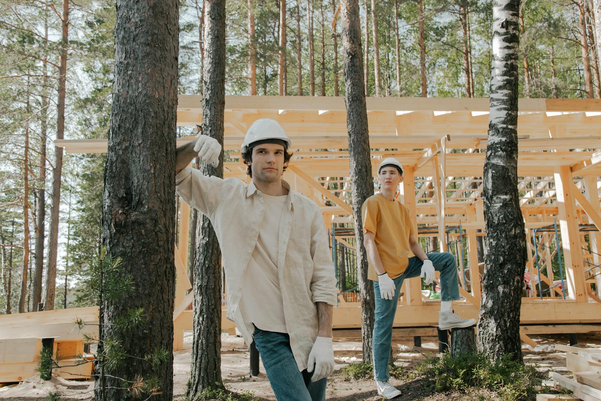 Two construction workers in protective gear at a forest site with wooden structure.